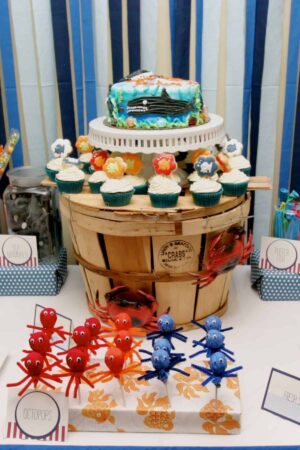 under the sea dessert and favor table with crepe paper backdrop and crab pot holding up a cake