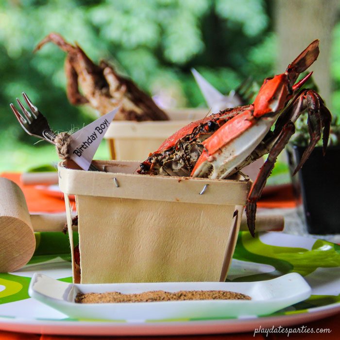 Crab feast place setting with a crab perched inside a berry basket, and a cocktail fork with a mini flag that says "Birthday Boy"