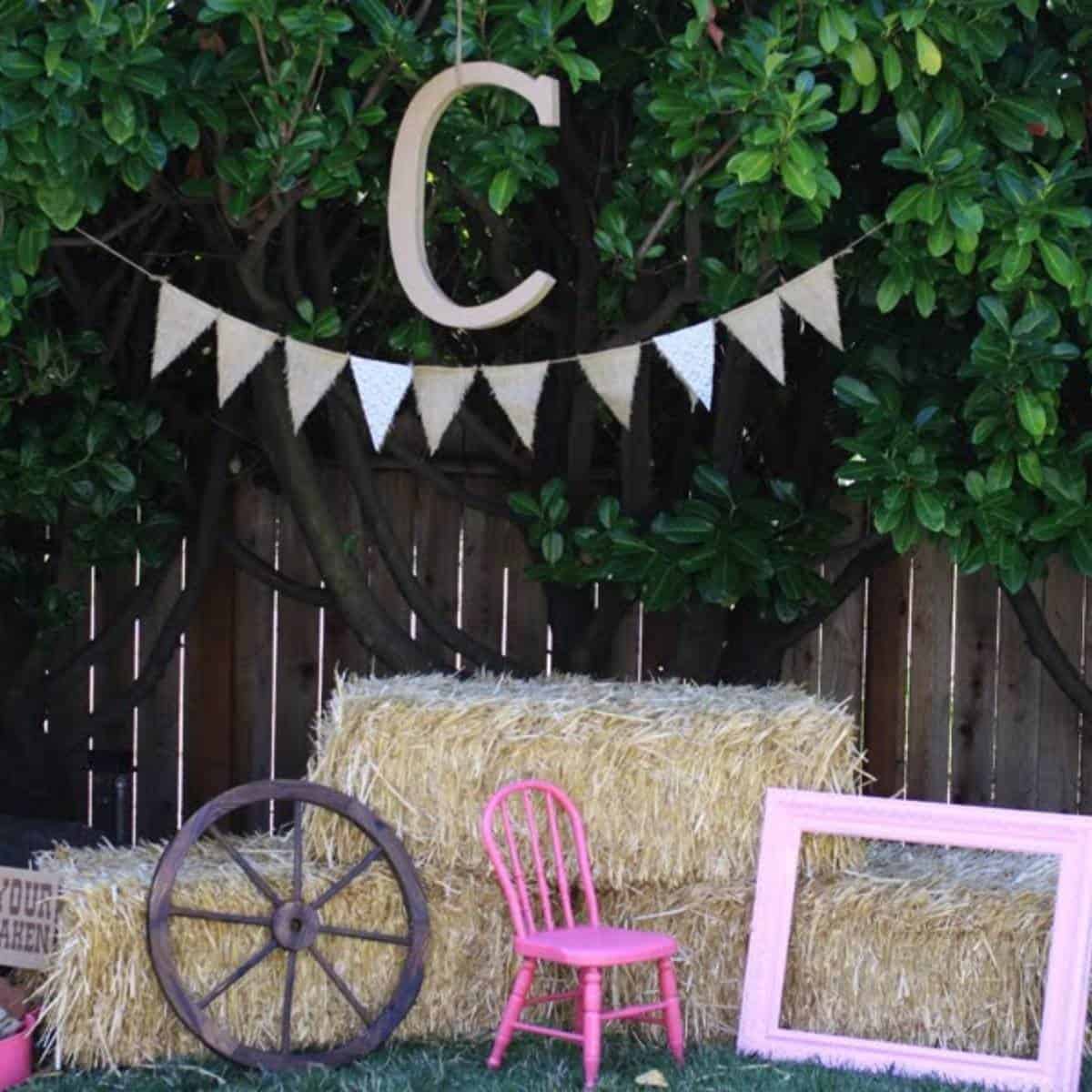 Photo booth setup with hay bales, a pink picture frame, a wagon wheel, and burlap bunting hanging from the tree above