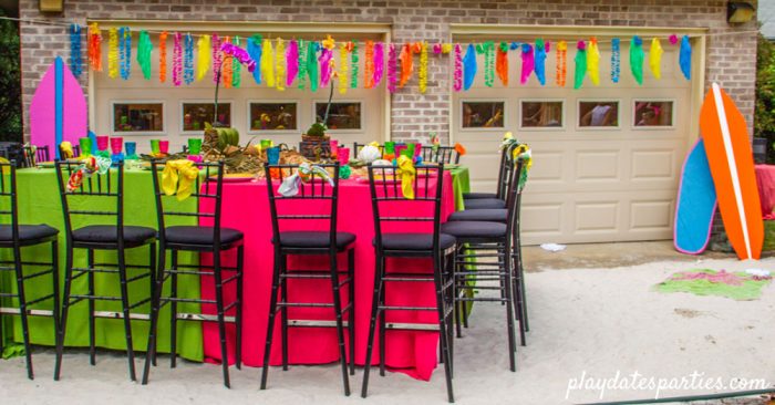 A temporary beach for a luau party with tables covered in colorful linens, a banner of grass skirts, and surf boards