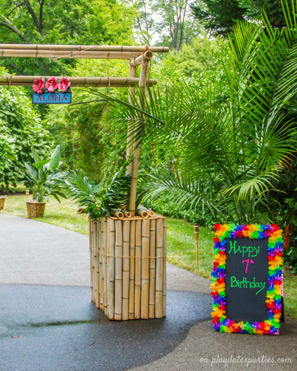 The entrance to a luau party, with a tiki arch, palm trees, and a happy birthday sign bordered in colorful Hawaiian leis.