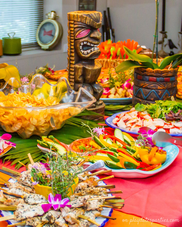 A buffet at a luau party with tiki centerpieces, vegetables, a bowl of chips, a vegetable tray and chicken skewers