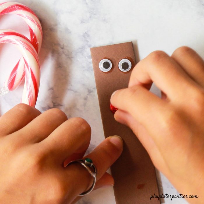 child's hands gluing a red jingle bell nose to a brown paper pocket with candy canes nearby