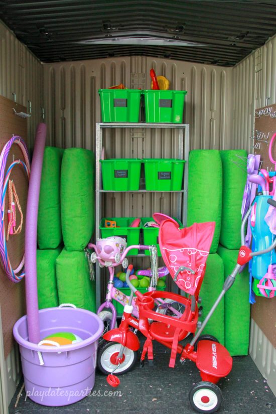 Outdoor toy storage shed completed with shelving, small toy boxes, hanging toys, a large bucket, a child's bicycle, and a tricycle.