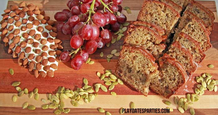 A pine cone cheese ball, banana bread and pepitas are served on a cutting board at a fall inspired brown and white baptism for a baby boy.