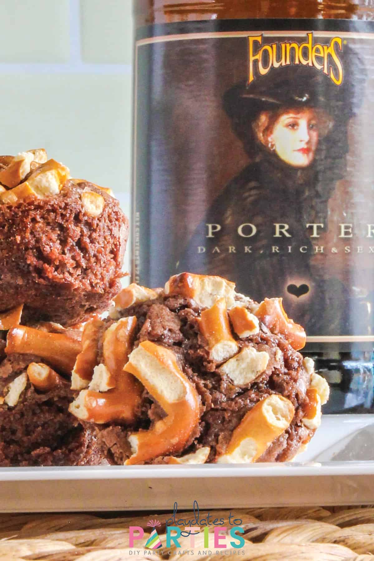 close up off a brownie bite with pretzel topping on a white plate in front of a bottle of beer