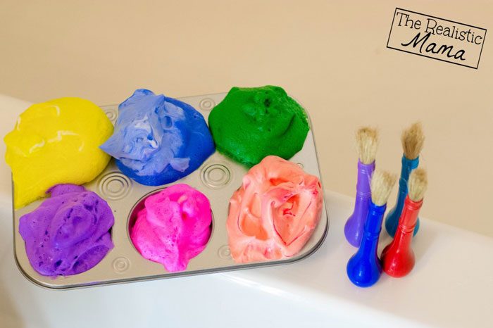 A muffin tin filled with colored shaving cream bath paint and four paint brushes next to it.