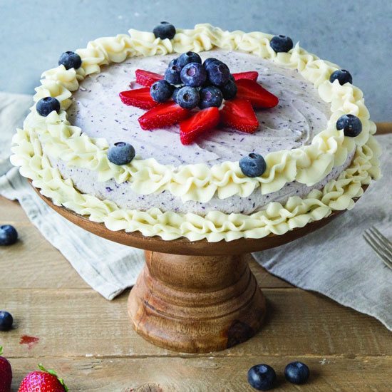 A blueberry ice cream cake with fresh berries, and piped frosting around the edges on a wooden pedestal cake stand.