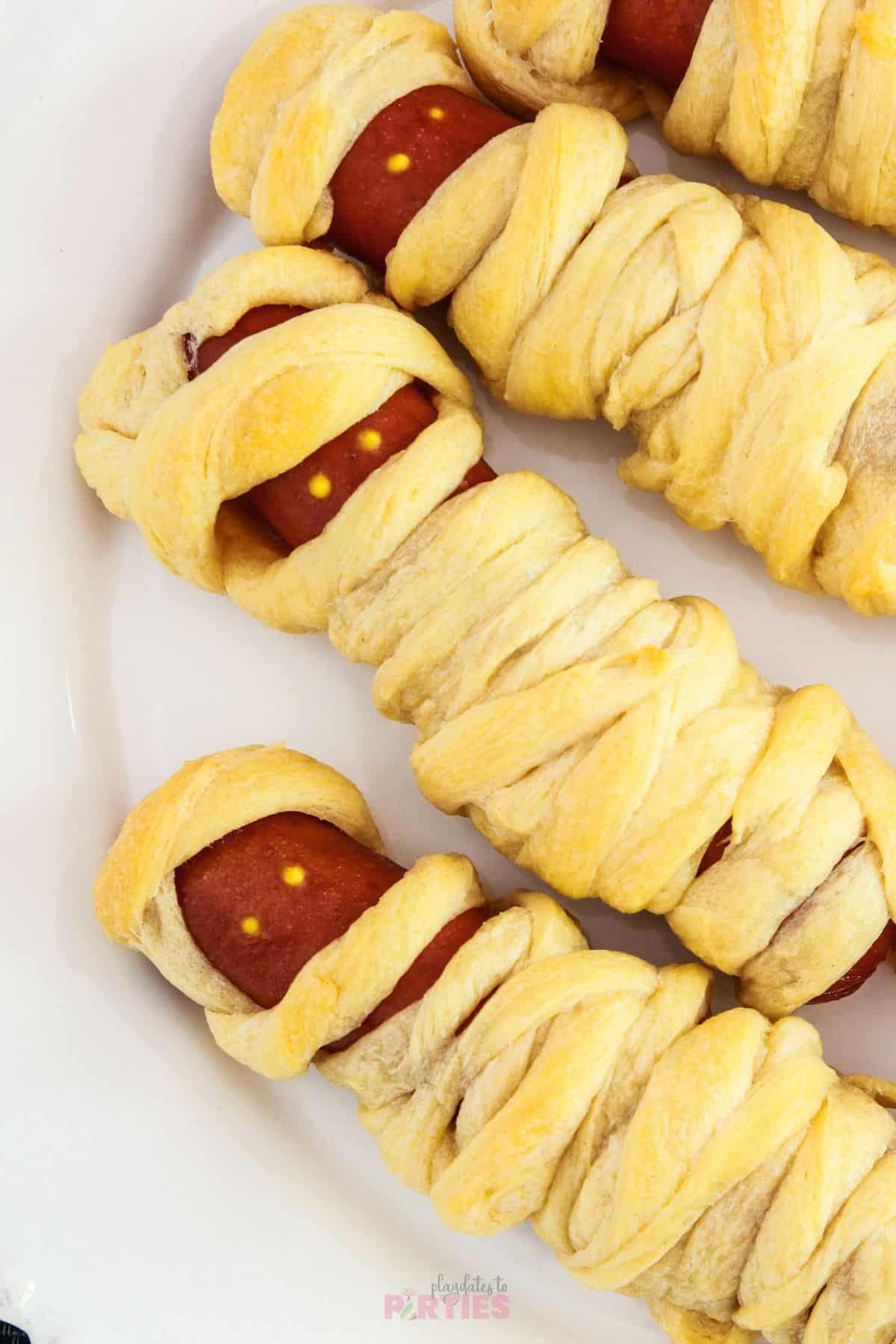 Cooked mummy dogs on a white plate with edible eyes.