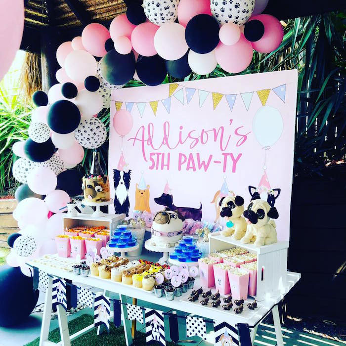 a puppy themed birthday party set up in pink and black with a backdrop that says "Addison's 5th paw-ty" and an asymmetrical pink, black and white balloon garland