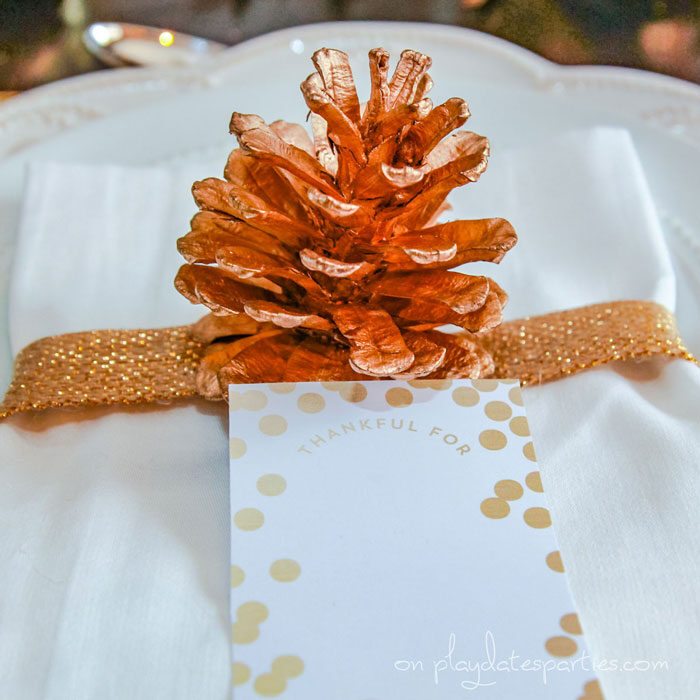 Close up of a napkin wrapped in gold ribbon with a gold pine cone and a card that says Thankful for