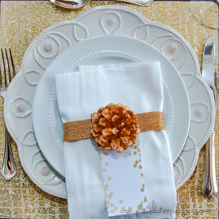 Overhead view of gold Thanksgiving table setting with a gold placemat, gold wrapped napkin, and gold pine cone