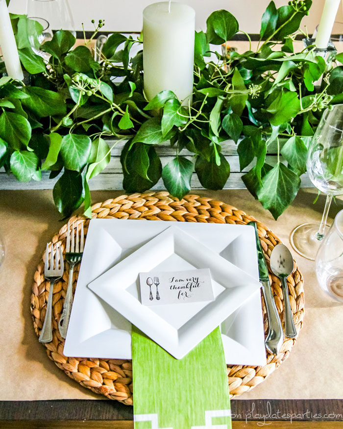 Thanksgiving table with a centerpiece that has a wood box filled with greenery, candles, woven placemats, white plates and a printable thankful card