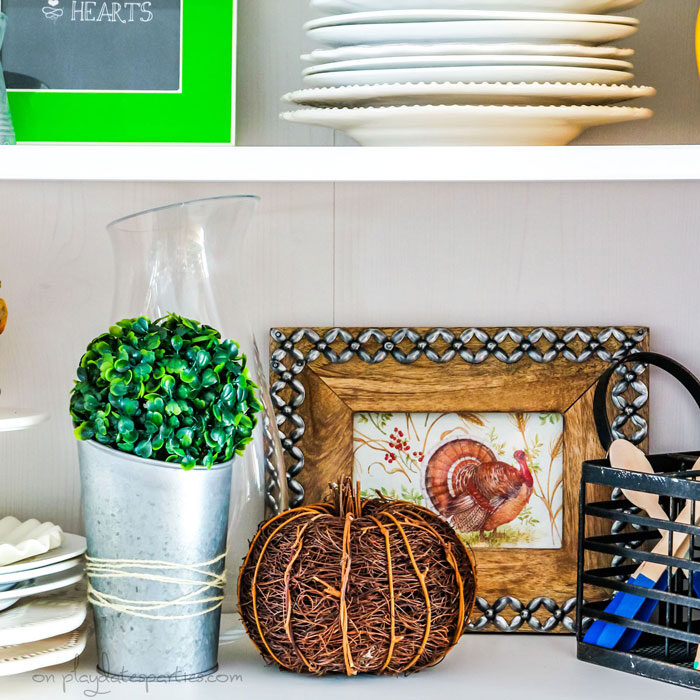 A kitchen hutch with inexpensive Thanksgiving decor: a napkin with a picture of a turkey that's been put inside a wood frame and a twig pumpkin