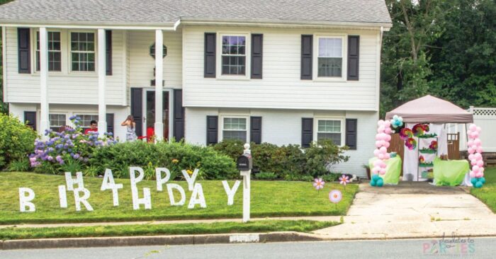 photo of a house with giant yard letters spelling happy birthday and a tent in the driveway with flower decorations