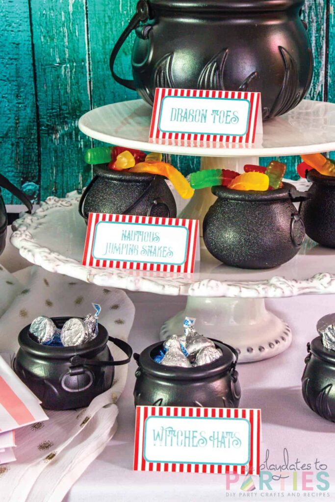 assorted candies in mini plastic cauldrons with food labels in front saying dragon toes, nautious jumping snakes, and witches hats