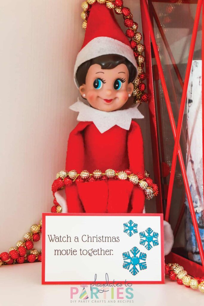 an elf sitting on a white shelf, tied up with a beaded garland with a card sitting in front that says watch a Christmas movie together