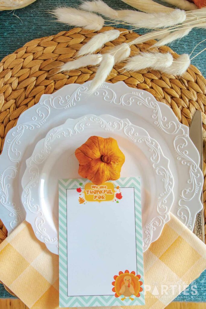 white plates on a woven hyacinth placemats with a miniature pumpkin and a we are thankful card placed on top