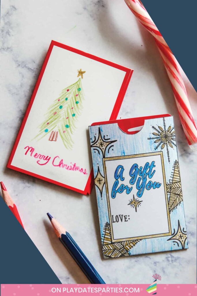 two color-in christmas gift card holders on a marble table with coloring pencils and a candy cane nearby