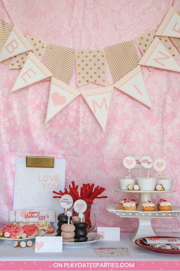 Valentine's day party setup with a banner that says "Be Mine", cupcakes, chocolate bars, mini donuts, treat bags, kisses, cookies, and a sign.