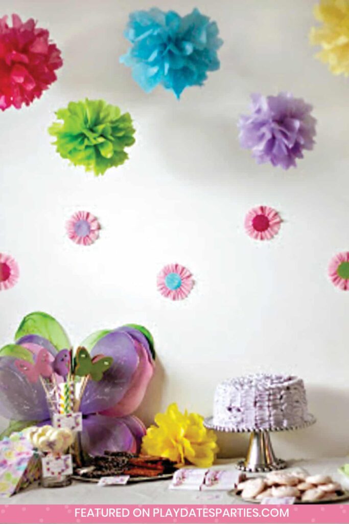 colorful tissue paper pom poms hanging over a dessert table with a cake, butterfly wings, cookies, and pretzel rods