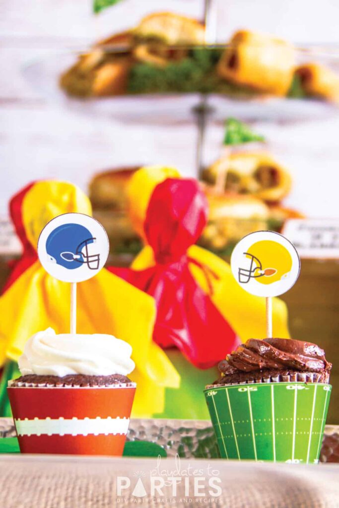 close up of two cupcakes. one has a printable topper with a blue football helmet the other has a printable topper with a yellow football helmet