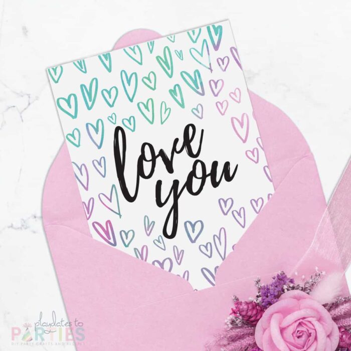 Picture of a 5x7 card peeking out of a pink envelope. The card has watercolor hearts surrounding the words love you in black script