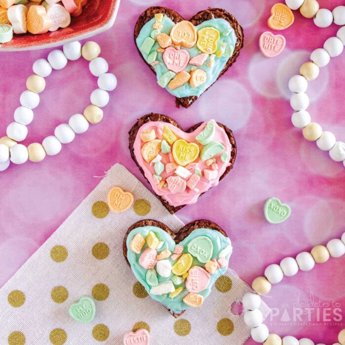 Three Valentine's Day brownies on a pink surface surrounded by party decorations and conversation hearts