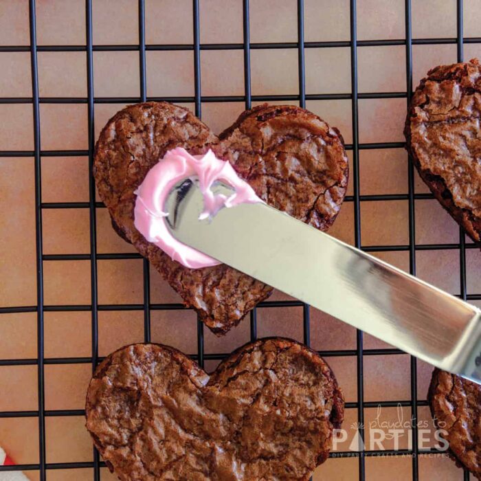 heart shaped brownies on a cooling rack with pink frosting being spread on one