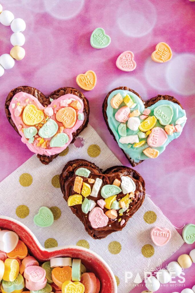 Three heart shaped brownies on a pink surface with pink, blue, and chocolate frosting and crushed conversation hearts on top.