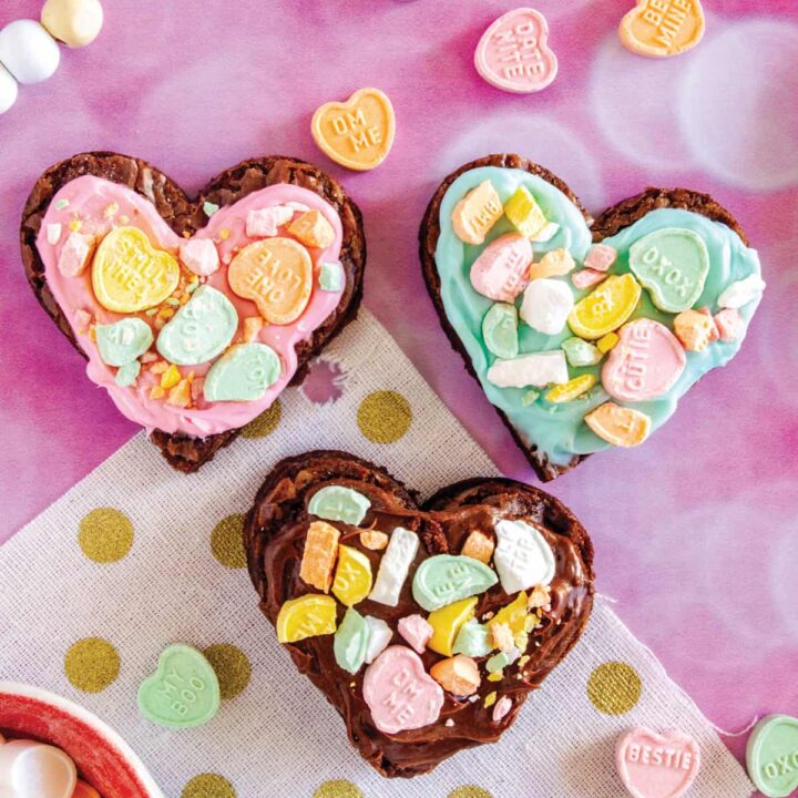 three heart shaped brownies on a pink surface with colorful frosting and conversation hearts