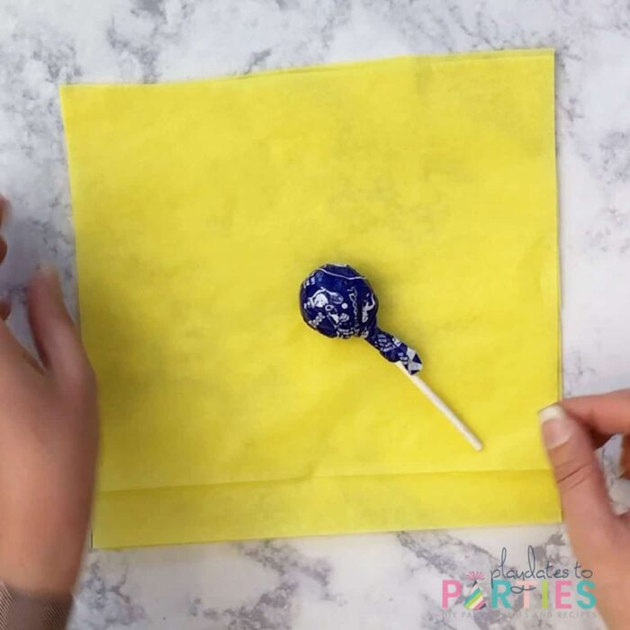 tootsie roll pop laying in the middle of square yellow tissue paper on a marble surface