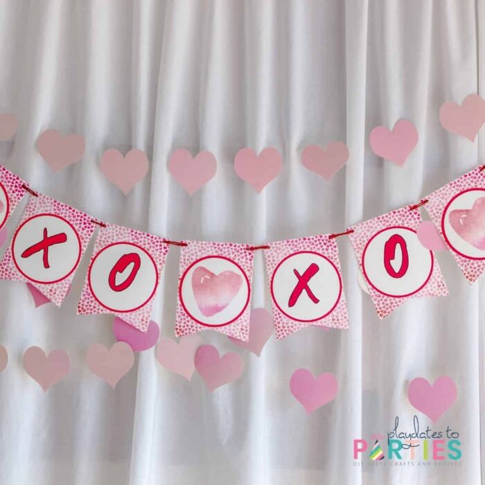 close up of a pink party banner with the letters XOXO in front of a white fabric backdrop and a garland made out of paper hearts.