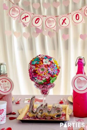 dining table set up for Valentine's Day with a centerpiece that says love, pink drink bottles, and a banner in the background that says XOXO