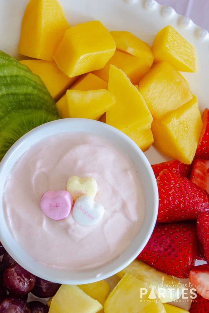 plate of strawberries, pineapple, grapes, and mango with a small bowl of pink dip in the center garnished with 3 candy hearts