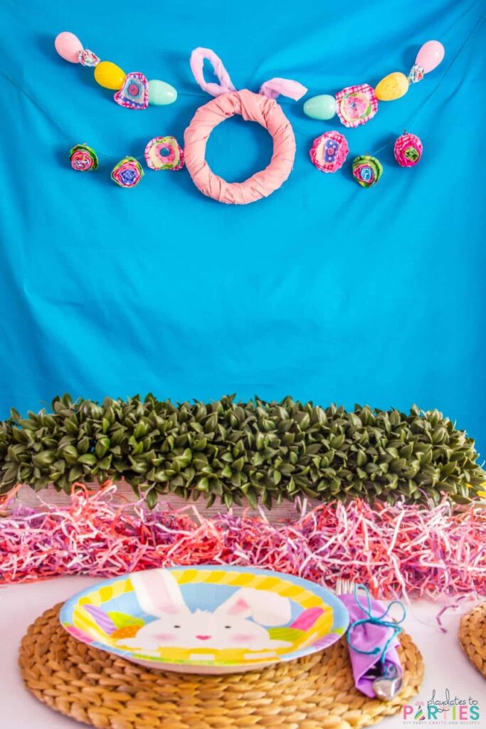 View of an Easter place setting and a bright blue backdrop with a bunny wreath and Easter egg garland