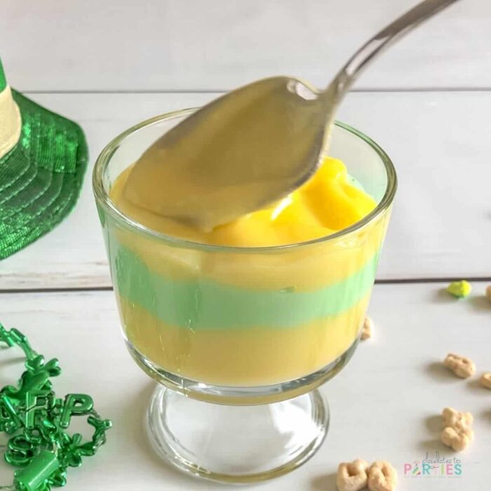 smoothing out the top layer of pudding in a parfait cup with St. Patrick's Day decorations and Lucky Charms cereal scattered around the cup