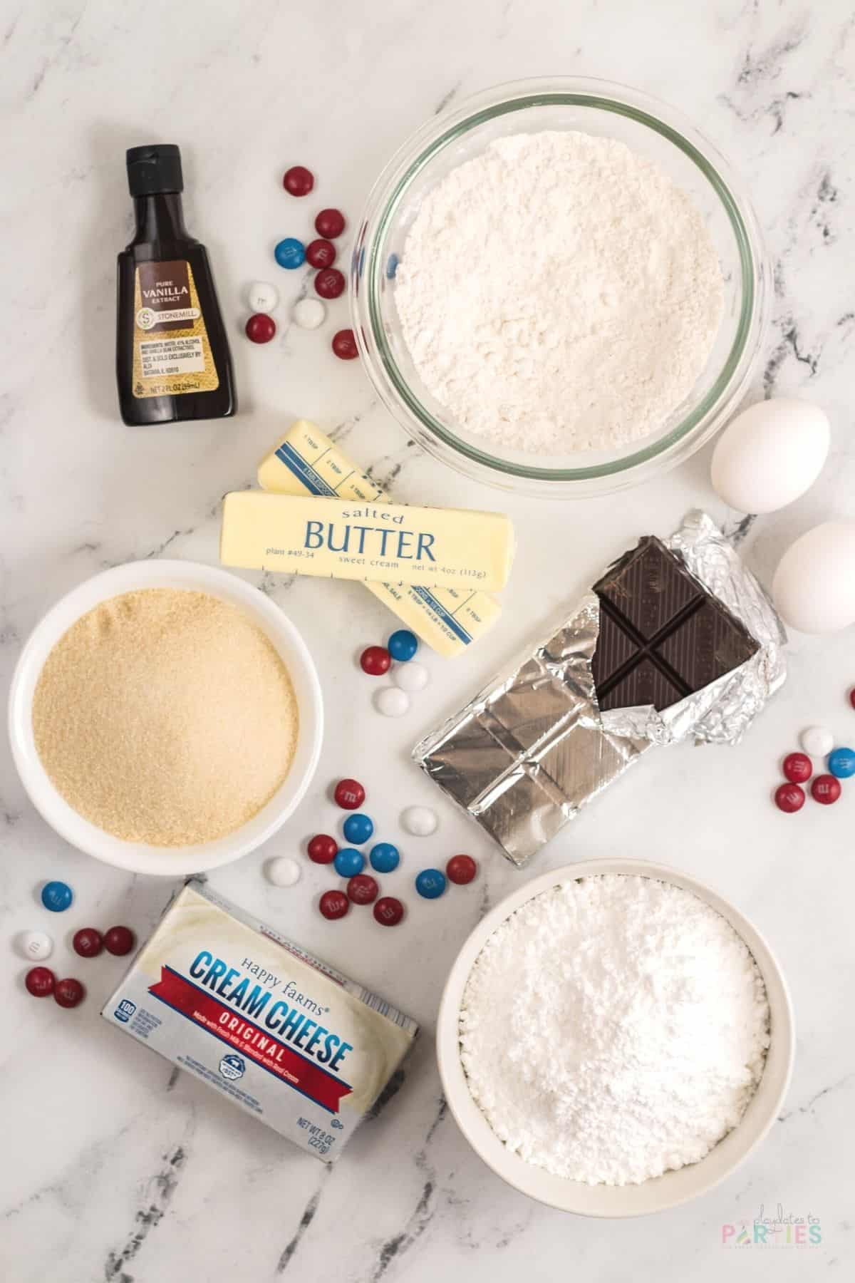 Ingredients for July 4th brownie pizza on a marble surface: vanilla extract, flour, eggs, butter, sugar, baking chocolate, cream cheese, powdered sugar, and candies