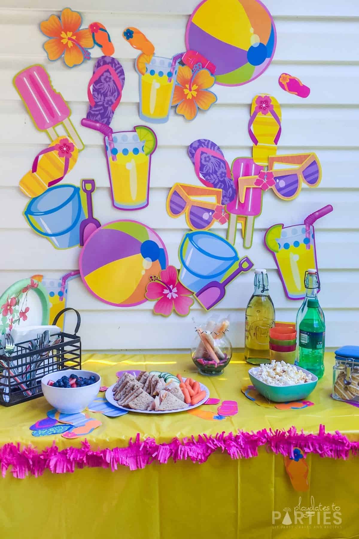 photo of a summer party table with beach themed paper cutouts on the wall, a yellow tablecloth and pink fringe around the edge