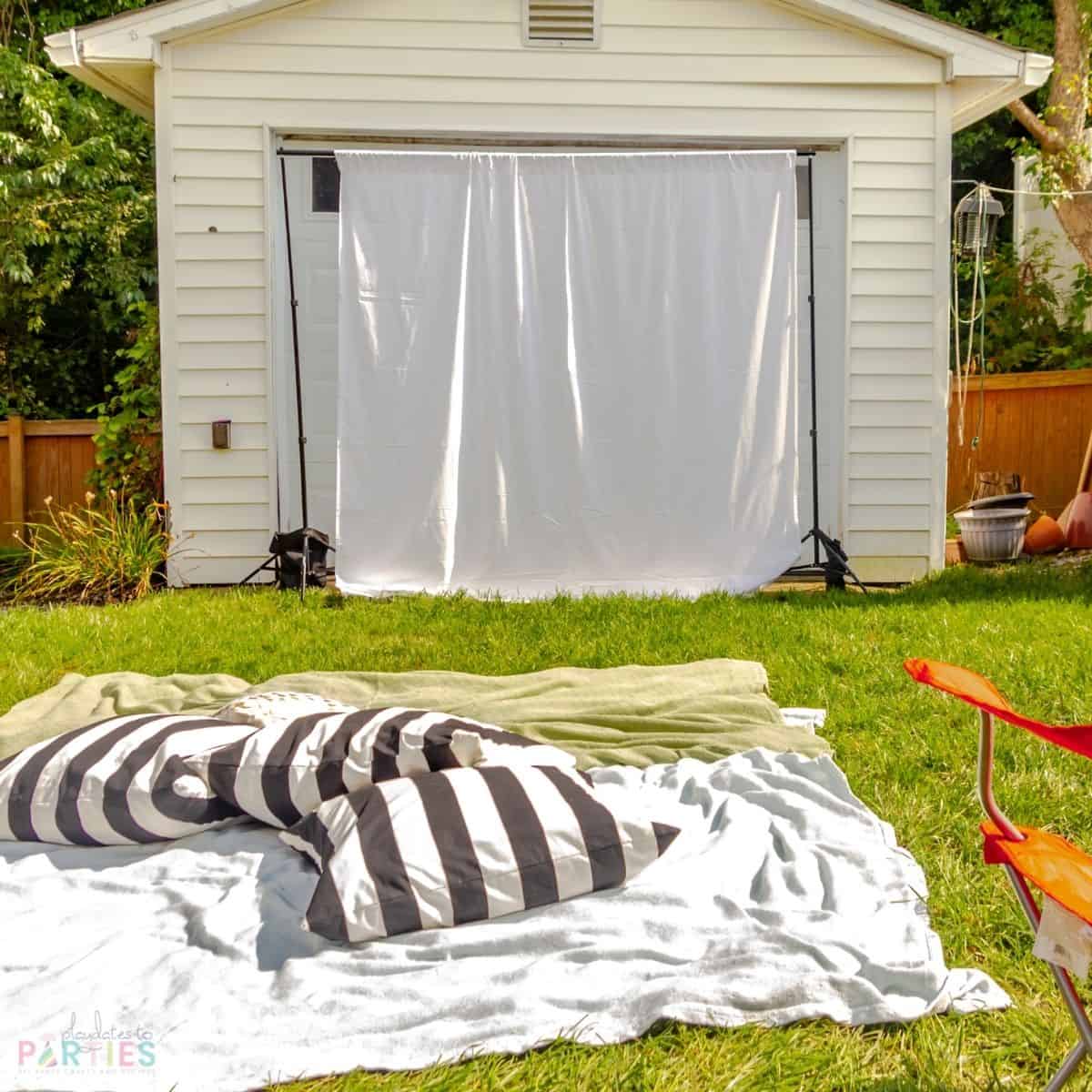 Simple outdoor movie setup with a party backdrop frame, and a white bedsheet. Throw blankets and house pillows provide a place for kids to hang out on the grass.