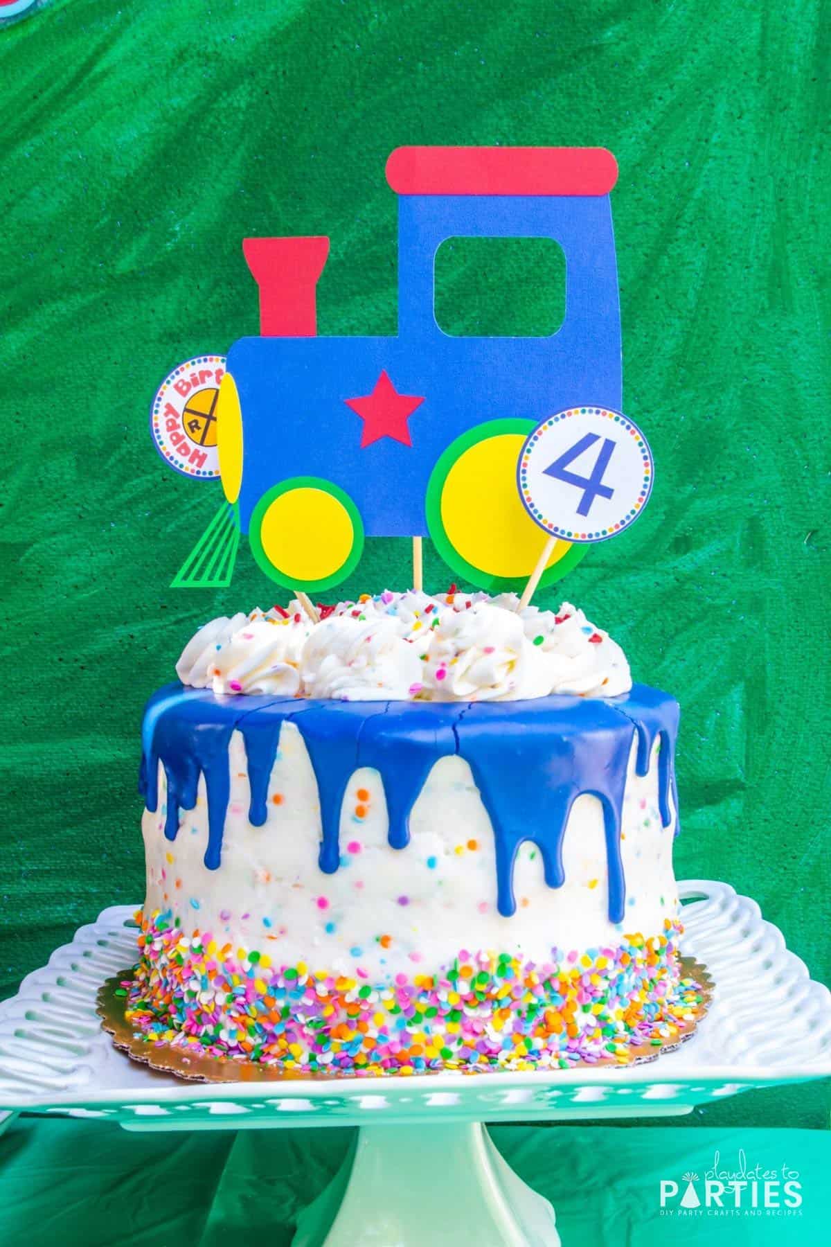 birthday party cake with blue drip icing, multicolored confetti sprinkles, and a paper train engine topper