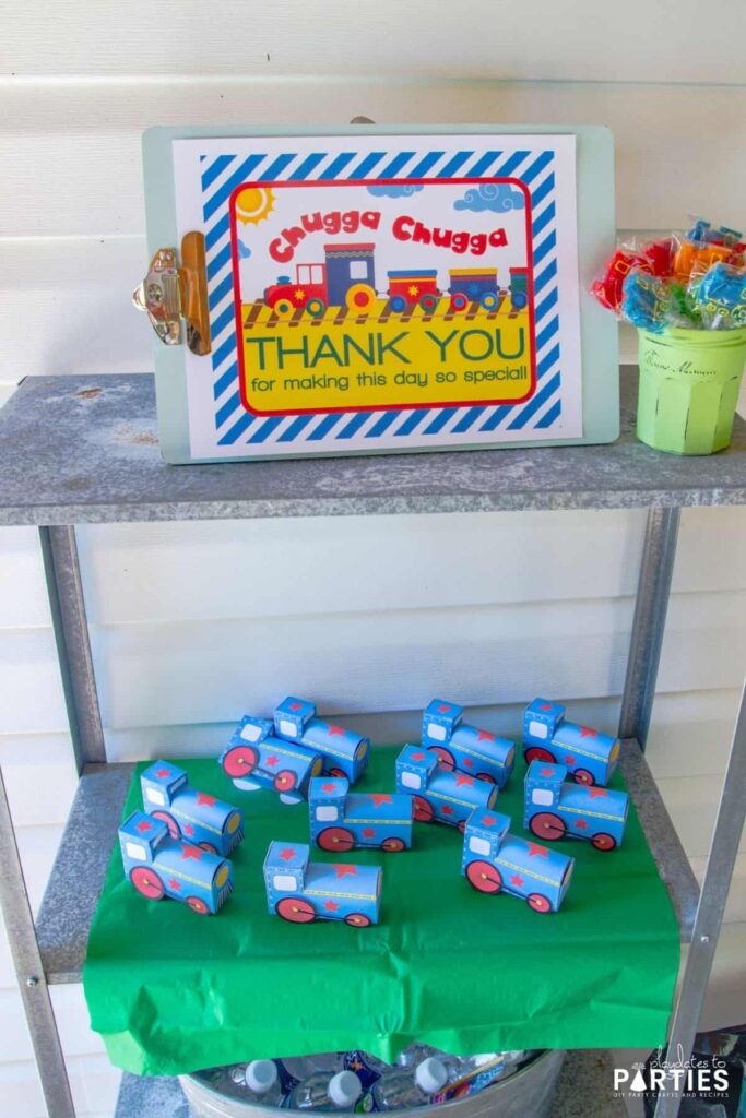 photo of outdoor shelves with mini trains as favor boxes, a jar of train lollipops, and a sign that says Chugga Chugga Thank You