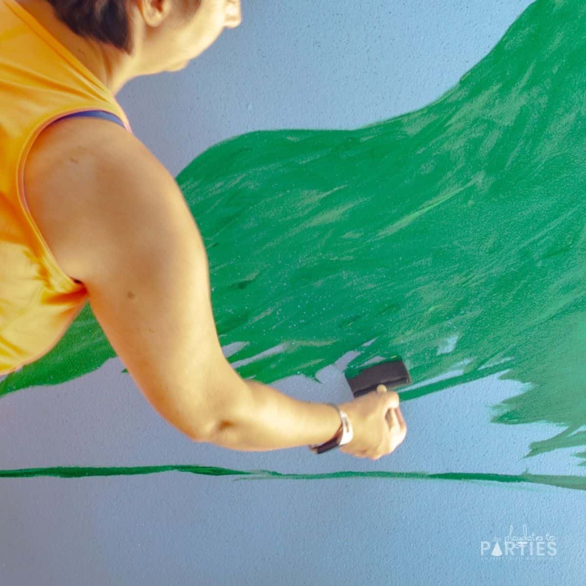 a woman painting a green grassy hill on a blue backdrop with a foam brush