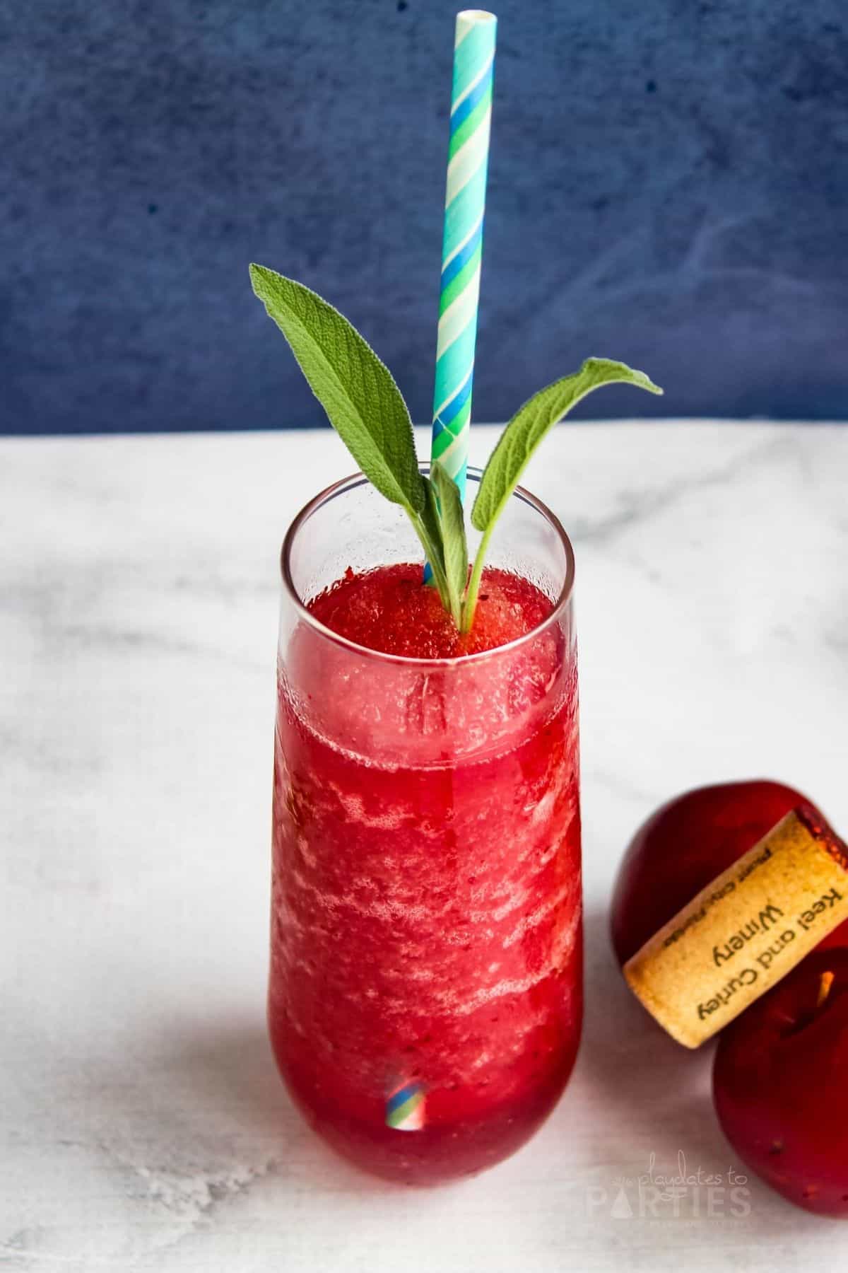 a bright red wine slushie in a stemless champagne flute garnished with basil and a colorful paper straw sitting next to fresh plums