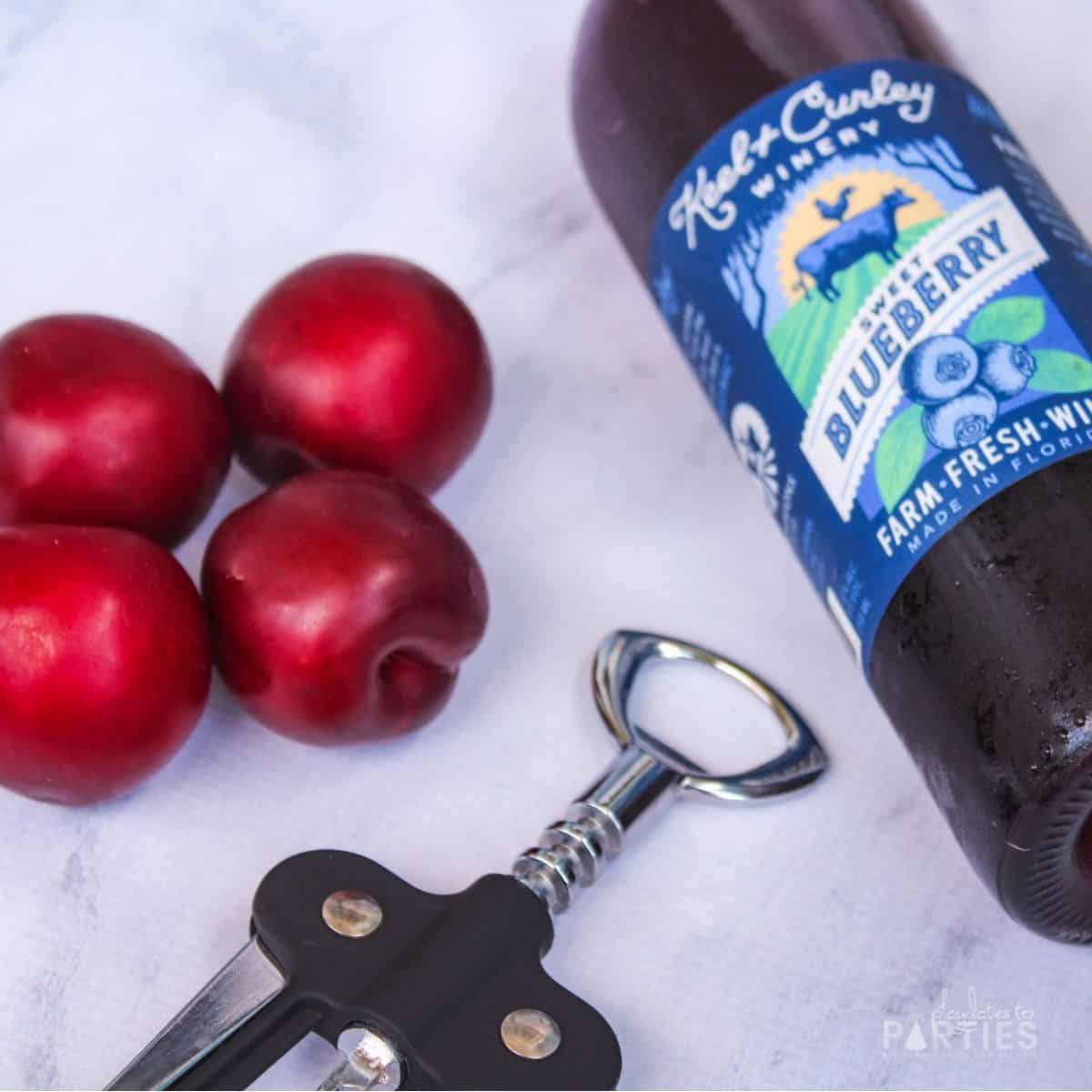 a bottle of wine and four plums on a marble surface next to a wine bottle opener