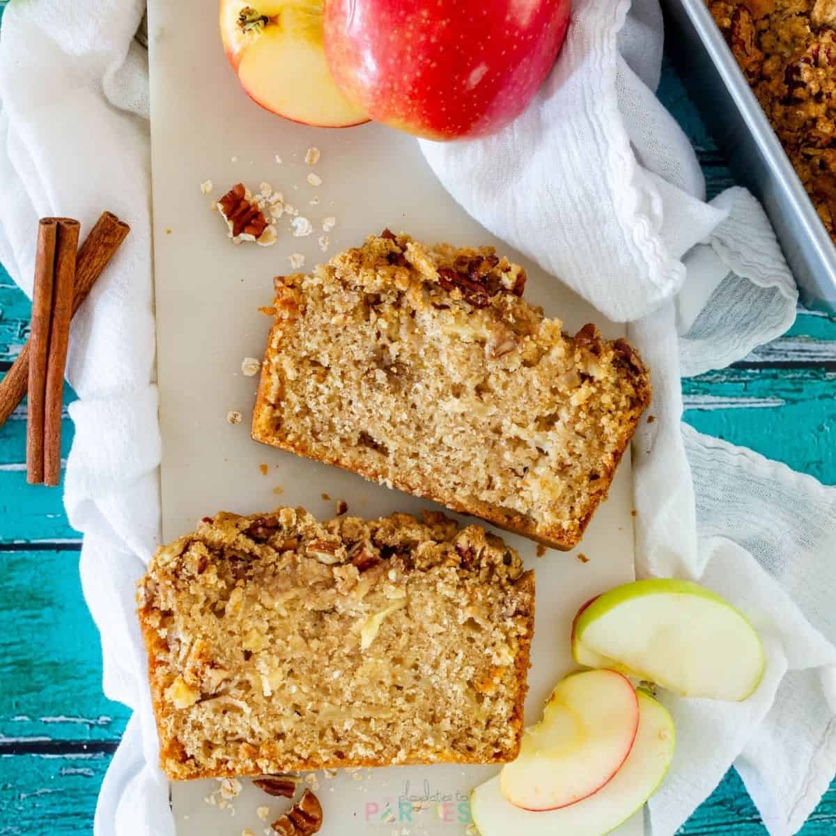 Two slices of apple bread on a marble board surrounded by apple slices, cinnamon sticks and a tea towel.