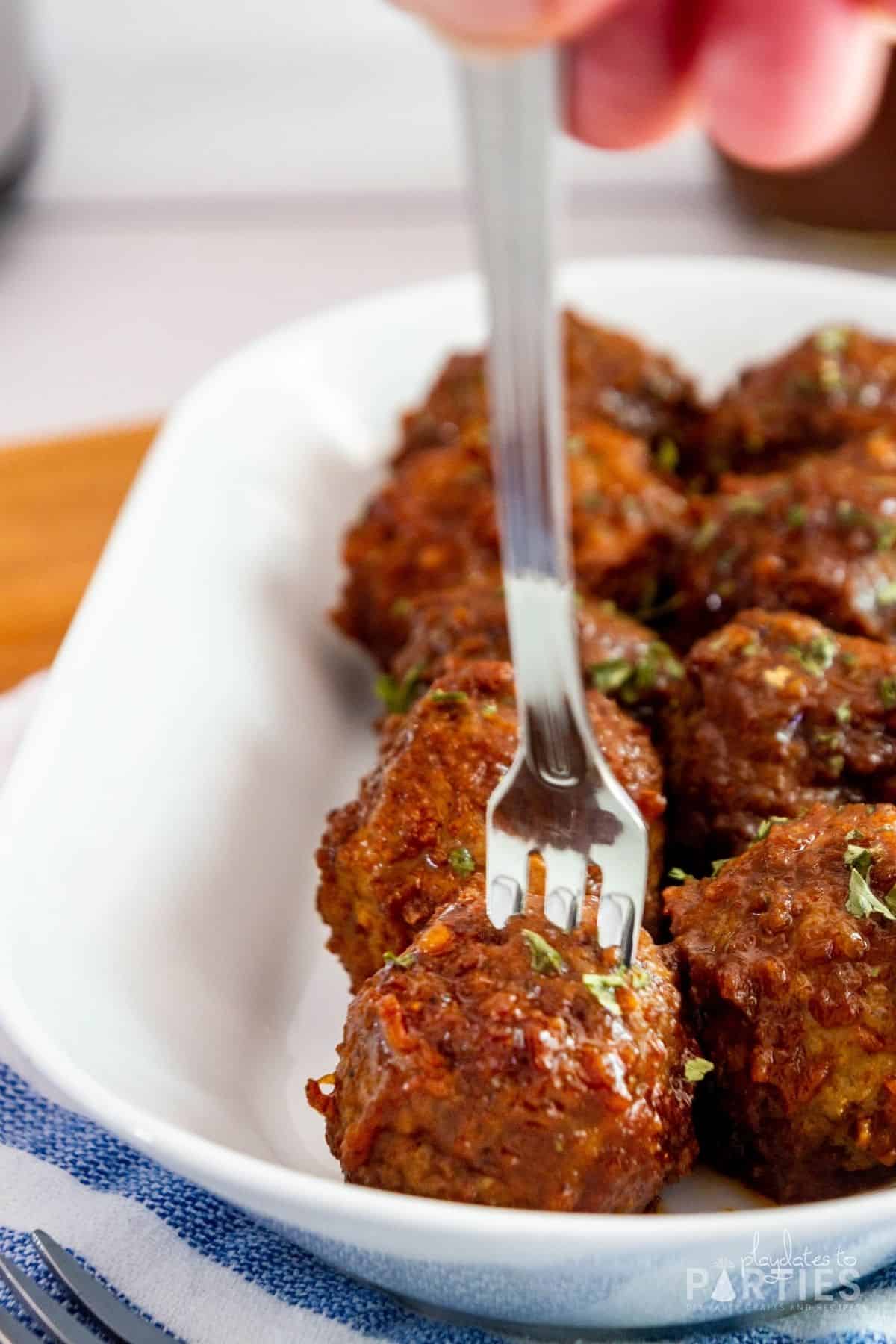 cocktail fork being pushed into a meatball on a white plate with a blue and white tea towel underneath