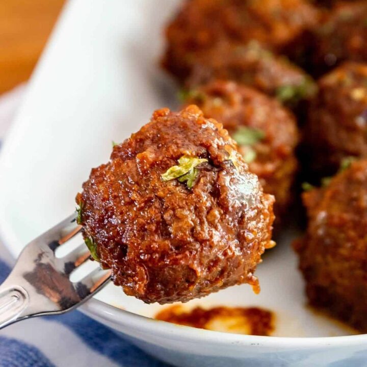 close up of a crock pot apple butter meatball on a cocktail fork with a plate of meatballs in the background