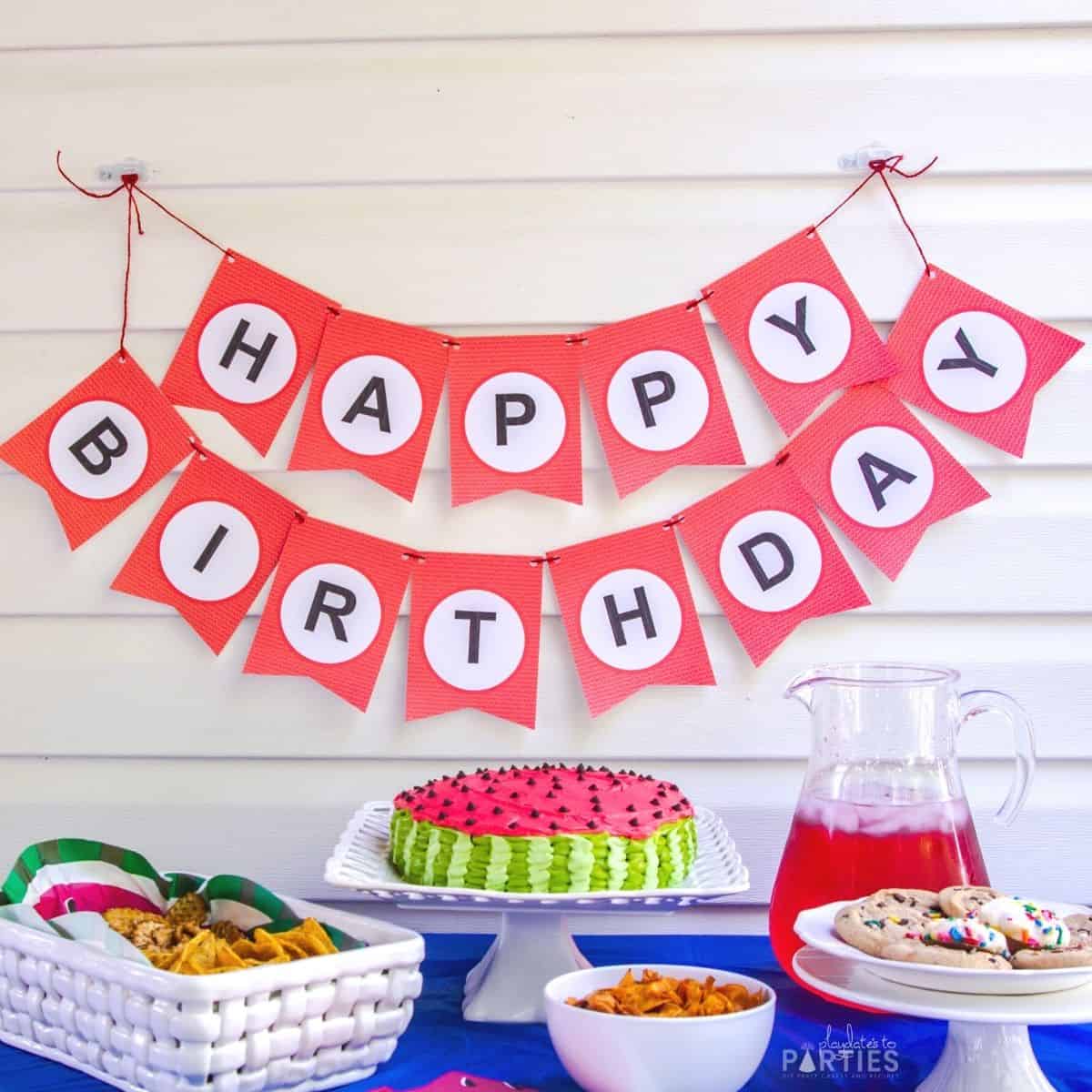 A red and white happy birthday banner behind a watermelon cake and snacks on a party table.
