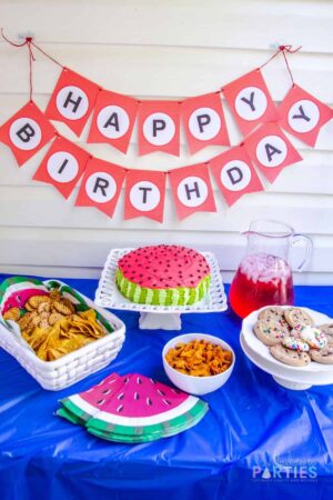 angled down photo of a party table and banner with birthday cake and snacks